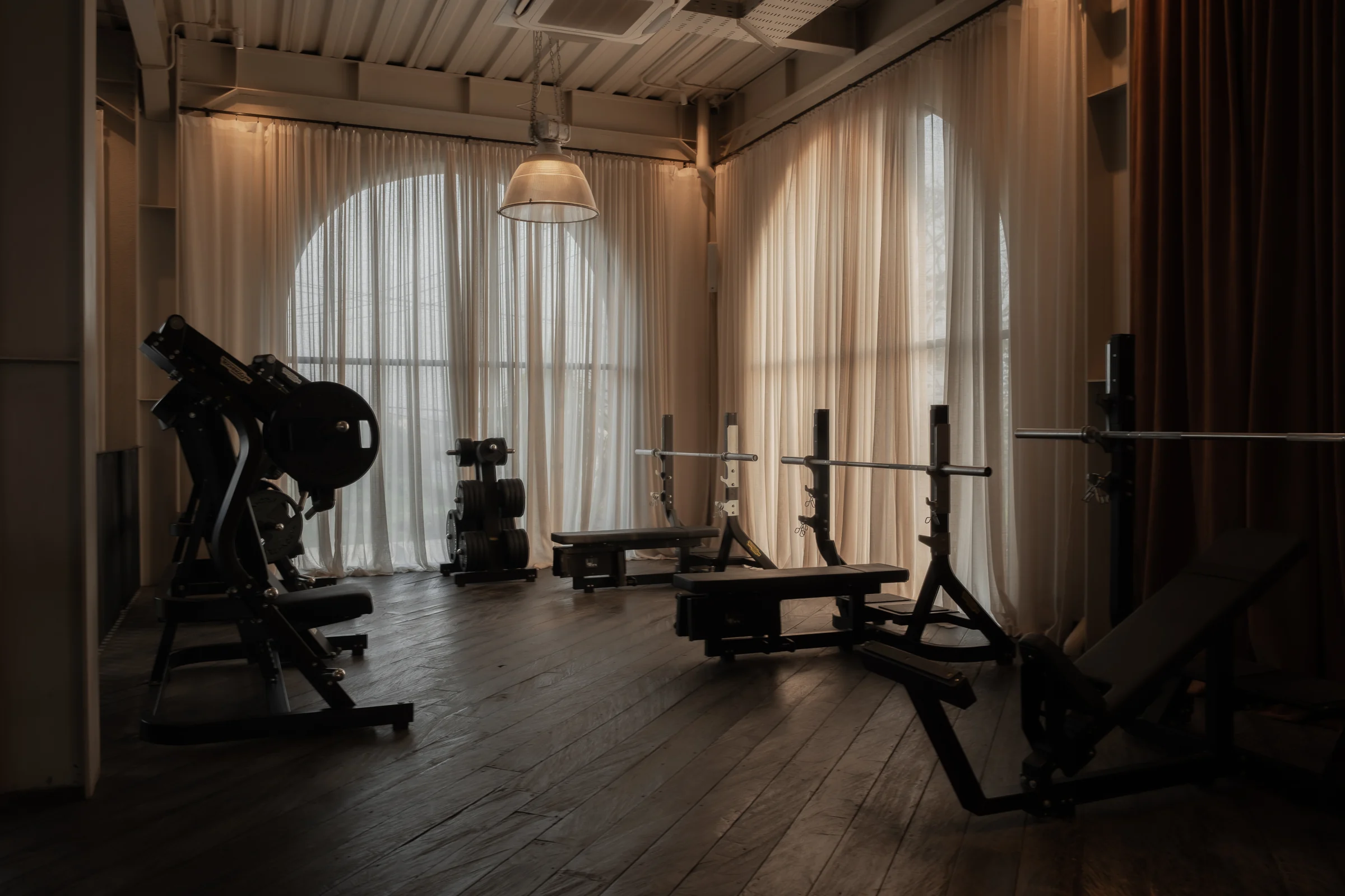 Bench stations and strength equipment beside tall, softly curtained windows.