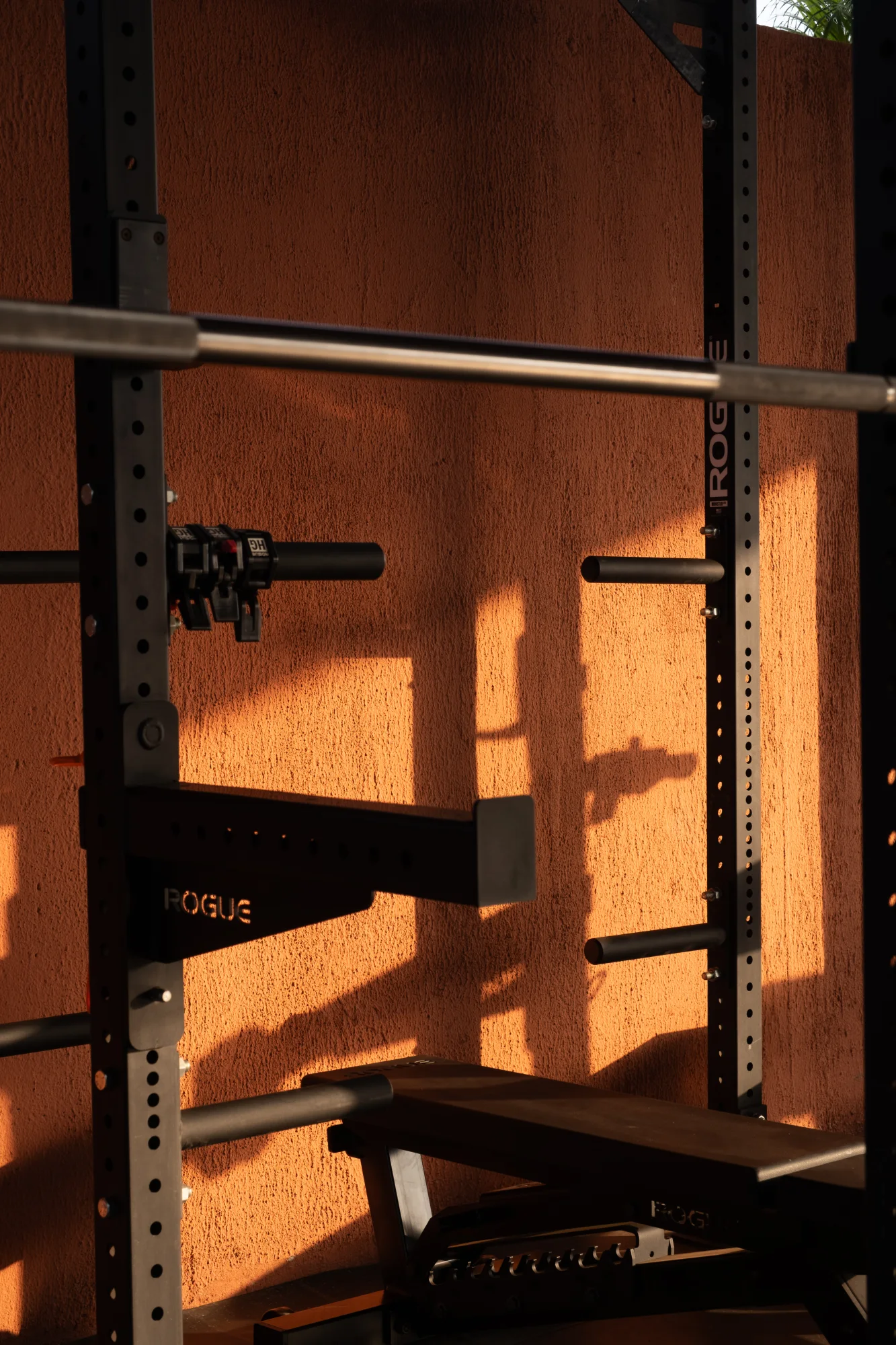 Sunlight and shadows across a squat rack and warm terracotta wall in the rooftop gym.