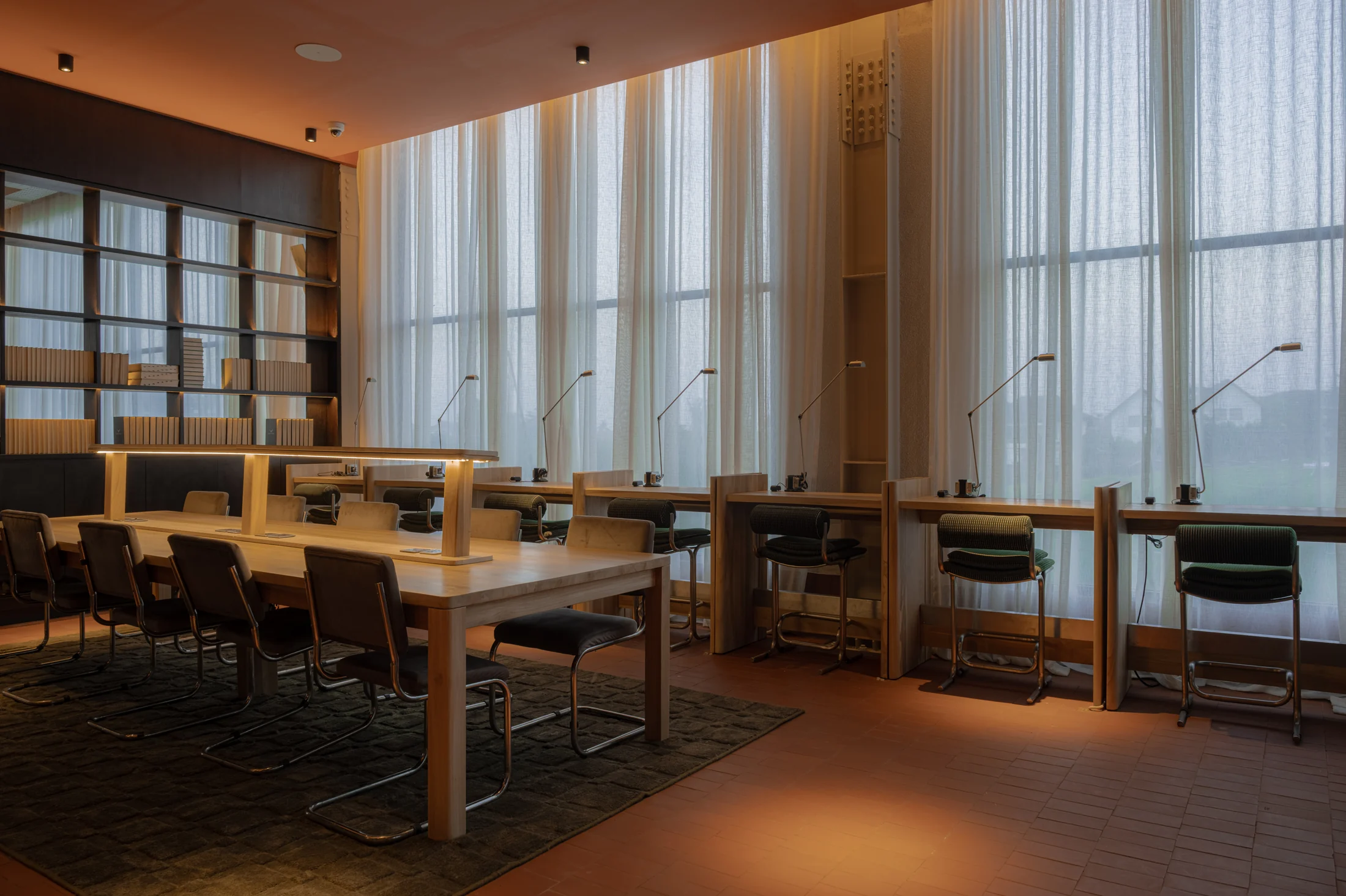 Shared timber tables and individual desks beside tall curtained windows in the coworking area.